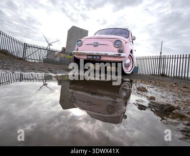 Der rosa Klassiker Fiat 500 spiegelt sich in einer großen Pfütze unter einem dramatischen bewölkten Himmel mit industriellen Elementen wie blyth northumberland uk wider Stockfoto