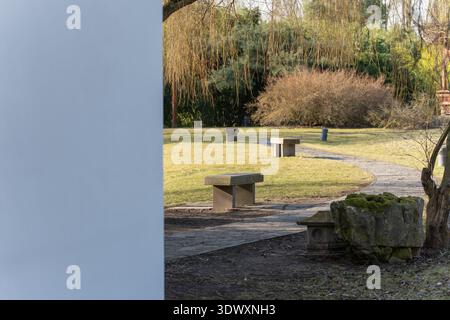 Ein geschwungener Steinweg führt durch einen ruhigen Garten mit Bänken. Sanfte Sonneneinstrahlung und Bäume schaffen eine ruhige Parklandschaft. Stockfoto