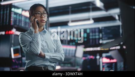 Young Asian Woman Trader steht im High-Tech-Büro und kommuniziert auf dem Smartphone und untersucht dabei die wechselnden Charts. Weiblich wechselt zwischen Bildschirmen, um Echtzeitindizes zu überprüfen. Expertenanalyse. Stockfoto