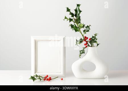 Stechpalmenzweige mit roten Beeren und grünen Blättern in Retro Donut Form Vase und leerer Vintage Holz Fotorahmen auf Glastisch vor Lichtwand. Tr Stockfoto