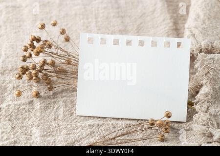 Leeres weißes Papier, Blatt oder Karte und getrockneter Leinpflanzenbund auf beigefarbenem Stoff aus reinem Leinen. Stockfoto