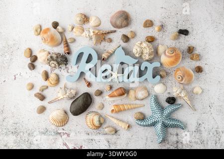 Hölzerne blaue Wort Relax, keramische Seesterne und verschiedene farbenfrohe natürliche Muscheln und Kieselsteine verstreut auf Grunge Stein Hintergrund. Sommerferien Stockfoto