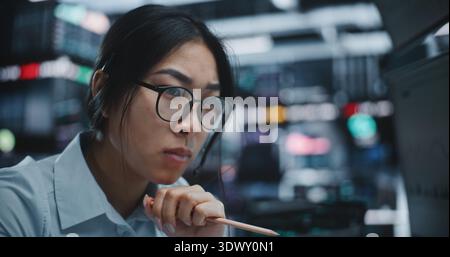 Stark fokussierte asiatische Traderin, die umfangreiche Marktdaten mit dem Bleistift in der Hand analysiert. Das Mädchen Wirbelt Nachdenklich Den Stift Zwischen Den Fingern. Strategische Analyse der digitalen High-Tech-Börse. Stockfoto