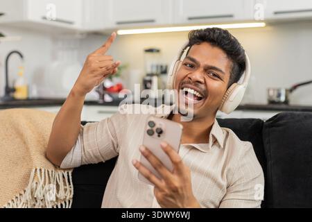 Indischer Mann zu Hause spielt Titel auf dem Smartphone mit schnurlosen Kopfhörern, die zu Beat und Lächeln nicken. Ein arabischer Kerl auf dem Sofa macht ein Rockschild und sendet Antworten im Chat, während Musik spielt und mit Spaß schlägt Stockfoto