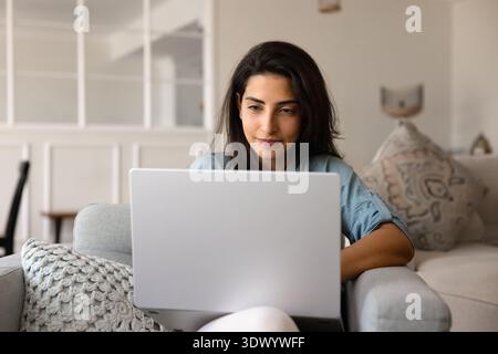 Die junge Latina sitzt bequem auf dem Sofa mit modernem Laptop Stockfoto