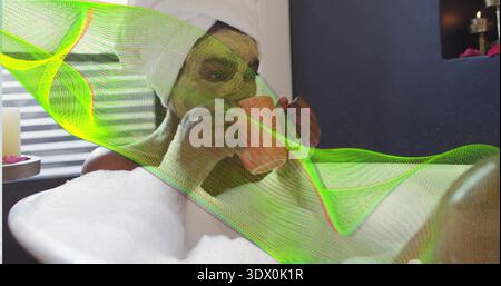 Liegend schwarze Frau schlürft Korallenbecher in der Hauswanne, trägt weißen Handtuch Turban, grüne Maske, Schaum Stockfoto