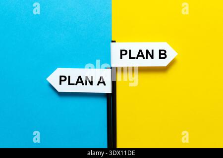 Ein Bild von zwei weißen Pfeilschildern mit der Bezeichnung PLAN A und PLAN B, die in entgegengesetzte Richtungen auf einem geteilten blauen und gelben Hintergrund zeigen und strategische Entscheidungen und Verfügbarkeitsplanung darstellen. Stockfoto