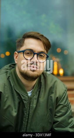 Vertikales Porträt eines attraktiven bärtigen kaukasischen Hipster-Typen mit Brille und grüner Jacke, der draußen vor einem dunklen Fenster posiert. Stockfoto
