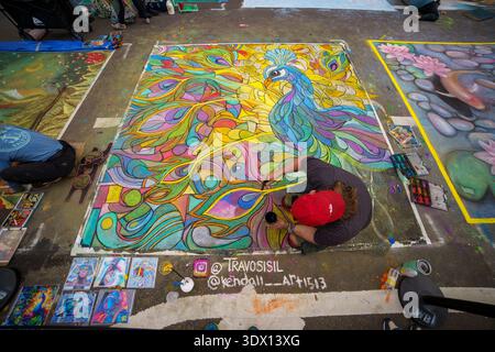 Ein männlicher Künstler sitzt auf der Straße und zeichnet beim Lake Worth Beach Painting Festival ein lebendiges, farbenfrohes Pfauengemälde. Stockfoto