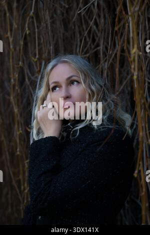 Nahporträt einer nachdenklichen kaukasischen blonden Frau im schwarzen Mantel, die gegen eine dunkle Wand mit trockenen Weinstöcken blickt. Stockfoto