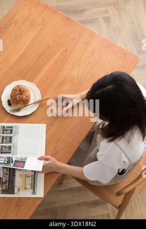 Frau, die sich am Holztisch mit Gebäck Kaffee und Magazin entspannt Stockfoto