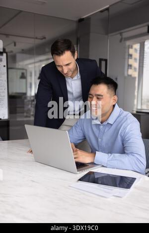 Lächelnder Geschäftsmann steht neben jungen männlichen Praktikanten, die am Notizbuch sitzen Stockfoto