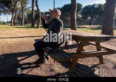 Mann sitzt auf einer Picknickbank in einem malerischen Wald und genießt einen Drink Wasser. Wanderausrüstung und helles Sonnenlicht unterstreichen die friedliche, erfrischende Atmosphäre im Freien Stockfoto