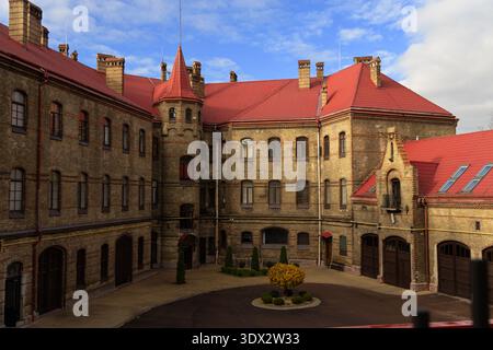 Ein großes, altes Backsteingebäude mit einem markanten roten Dach und einem charmanten Innenhof mit gepflegten Büschen und einem kleinen Baum Stockfoto