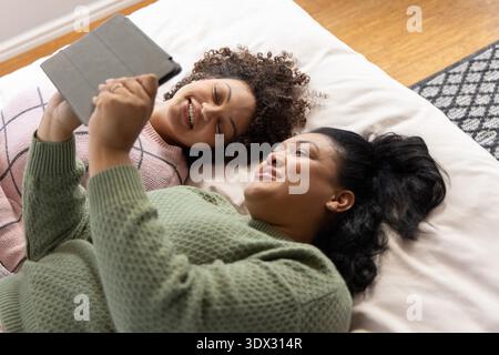 Verschiedene Freundinnen liegen nebeneinander auf dem Bett und halten ein Tablet mit Hülle in kuscheligen Pullis. Duo, Frauen, Schlafzimmer, Sonnenlicht, Freizeit, Snug, device || Model veröffentlicht Stockfoto