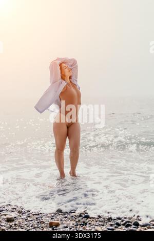 Frau Strand Sonnenaufgang Frau in Body und Hemd genießt morgens Meeresbrise am Kieselstrand mit friedlicher Sommerstimmung Stockfoto