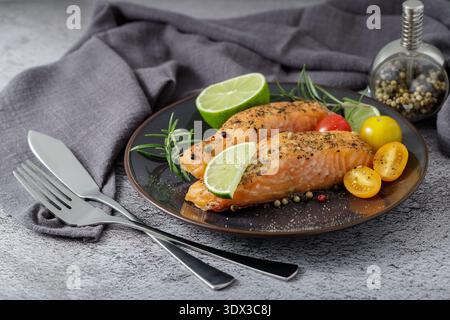 Gegrillte Lachssteaks mit Kräutern, Gemüse, Limette und Gewürzen auf einer Keramikplatte. Das Gericht wird auf einem Steintisch serviert Stockfoto