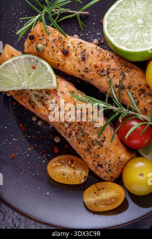 Zwei gegrillte Lachssteaks mit Kräutern, Tomaten, Limette und Gewürzen auf einer Keramikplatte. Steinhintergrund, Draufsicht Stockfoto