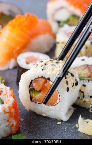 Sushi mit Avocado, Lachsfisch und Sesamsamen mit schwarzen Stäbchen auf dunklem Steinbrett. Stockfoto