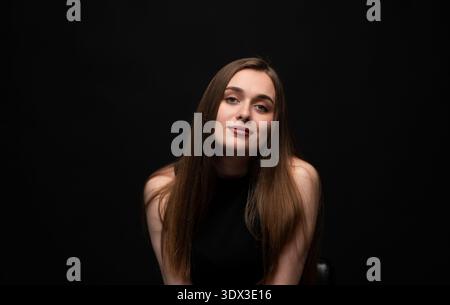 Junge Frau mit langen braunen Haaren, die selbstbewusst in einem Studio-Porträt mit dunklem Hintergrund posiert Stockfoto