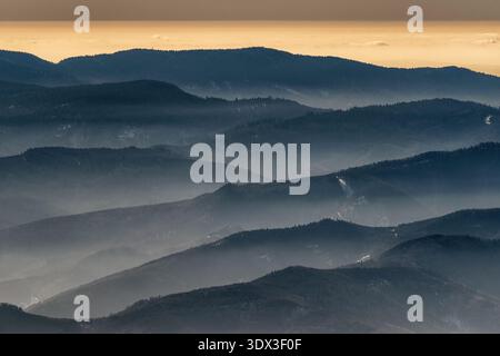 Mehrschichtige Bergkämme der Niederen Tatra in der Slowakei bedeckt mit winterlichem Dunst bei Sonnenuntergang mit weichem goldenem Licht und atmosphärischer Tiefe. Stockfoto