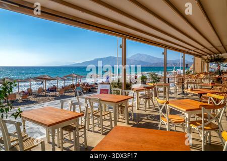 Kalamaki, Griechenland - 4. September 2025: Terrasse des Strandrestaurants mit Holztischen und -Stühlen mit Blick auf Sonnenliegen und das Libysche Meer an der Südküste Stockfoto
