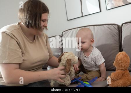 Frau und Baby sitzen auf der Couch und spielen mit gefülltem Hund und Teddybär. Fröhliche Mutter-Kind-Bindung und Elterninteraktion für Familienmomente am Muttertag. Fotografie Stockfoto