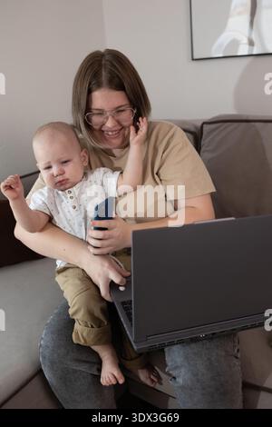 Frau, die das Baby auf dem Schoß hält, während sie im gemütlichen Wohnzimmer am Laptop arbeitet. Multitasking-Mutter, die Fernarbeit und Kinderbetreuungsaufgaben in Einklang bringt. Eltern- und Home-Office-Konzept für Muttertagsfeiern. Stockfoto