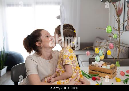 Eine lächelnde Mutter und ihre Tochter, die Hasenohren tragen, teilen sich einen zarten Kuss in ihrem Haus. Stockfoto