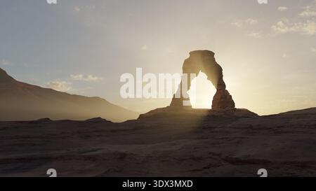 Die Sonne geht hinter Delicate Arch im Arches National Park auf. Das Sonnenlicht scheint durch den Bogen und schafft eine helle Kontur. Berge und Wolken sind sichtbar Stockfoto