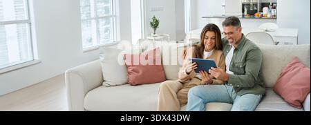 Glückliches Paar mittleren Alters mit einem digitalen Tablet, das sich zu Hause auf dem Sofa entspannt. Stockfoto
