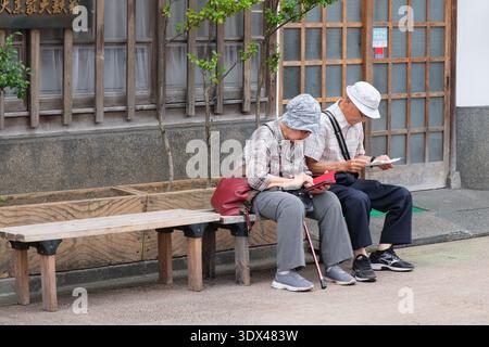 Tokio, Japan - 07. August 2025: Seniorenpaar in lässiger Kleidung, das Telefon auf einer Holzbank in einer traditionellen japanischen Straße mit hölzernem Archit überprüft Stockfoto