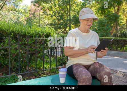 Eine ältere kaukasische Frau liest ein E-Book auf einer Parkbank. Stockfoto