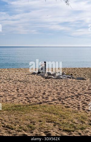 Eine Person sitzt auf einem Steinhaufen an einem Sandstrand, während sie mit einem Smartphone ein Bild des ruhigen Ozeans macht. Stockfoto