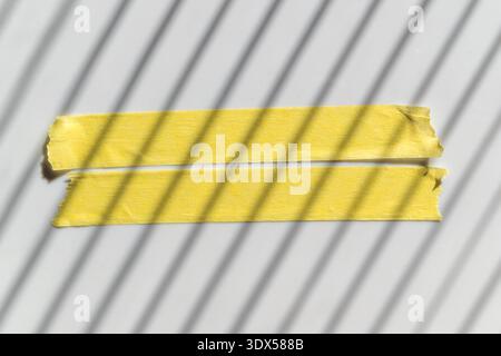 Abgebrochener gelber Kreppstreifen auf hellem Hintergrund mit dramatischen gestreiften Schatten, minimaler abstrakter Textur und grafischer Beleuchtung. Draufsicht. Stockfoto