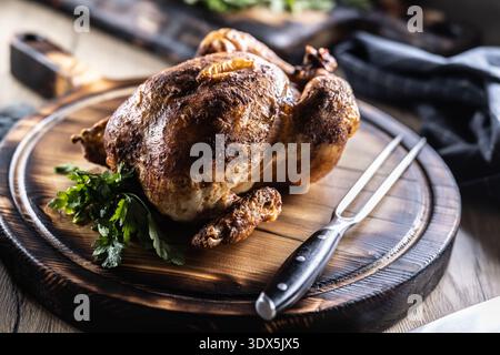 Ein ganzes, goldbraun gebackenes Hühnchen wird auf einem Holzschneidebrett serviert, garniert mit frischer Petersilie und begleitet von einer Fleischgabel in einem Rusti Stockfoto