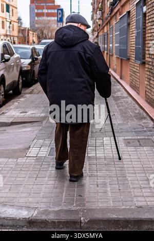 Badalona, Spanien - 3. Februar 2026: Rückansicht eines älteren Mannes mit flacher Mütze und Wintermantel, der mit einem Stock auf einem gekachelten Bürgersteig geht, illustriert Stockfoto