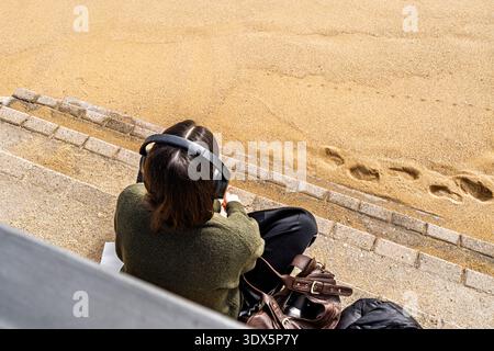Blick über der Decke auf eine nicht erkennbare junge Frau mit Kopfhörern, die auf Steinstufen am Sand sitzt und den Lebensstil digitaler Nomaden repräsentiert, Podcasts, Stockfoto