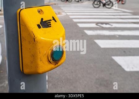 Nahaufnahme des gelben Druckknopfes für Fußgänger am Pfosten mit verschwommenem Zebraübergang und Radfahrern im Hintergrund, städtisches Verkehrssicherheitskonzept Stockfoto