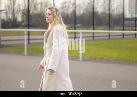 Seitenansicht einer Frau, die einen hellen Mantel trägt, während sie im Freien in einer Parkumgebung spaziert, aufgenommen bei natürlichem Tageslicht mit einer entspannten und ruhigen Stimmung. Stockfoto