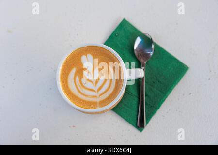 Cremiger Latte mit zarter Blattlattkunst, präsentiert in einer weißen Tasse auf einer grünen Serviette neben einem Löffel. Minimaler Overhead-Aufbau sorgt für gemütliche Morgenstunden Stockfoto