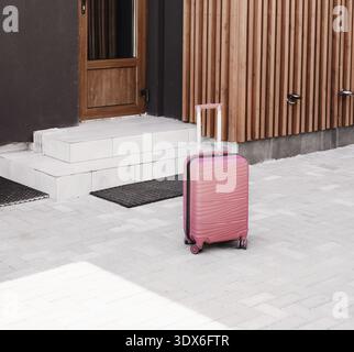Rosafarbener Gepäckkoffer, der vor dem modernen Hauseingang mit Holzverkleidung steht. Konzept von Reise, Tourismus, Urlaubsreise oder Umzug ins neue Stockfoto