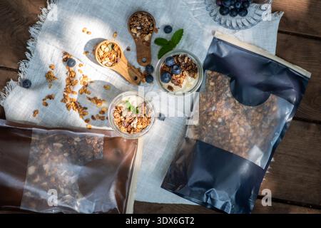 Blick von oben auf natürliches Müsli, serviert in Glasgläsern mit Joghurt und frischen Heidelbeeren auf einem Tisch. Holzlöffel und mit Müsli gefüllte Beutel fügen ein oder hinzu Stockfoto
