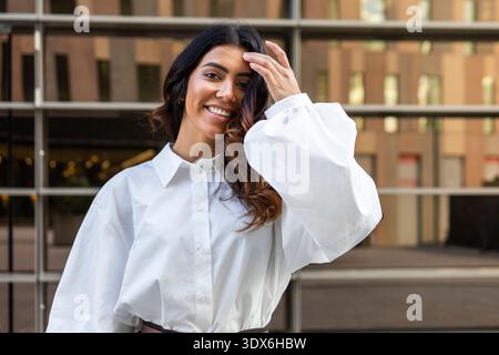 Hispanische Geschäftsfrau mit langen dunklen Haaren, die selbstbewusst in einer weißen Bluse lächelt und vor einem modernen Bürogebäude steht. Sie scheint Professi zu sein Stockfoto