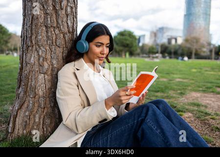 Eine hispanische Frau sitzt in einem Stadtpark an einem Baum, trägt Kopfhörer und liest ein Buch. Sie trägt Jeans und einen Blazer und umschließt einen Stockfoto