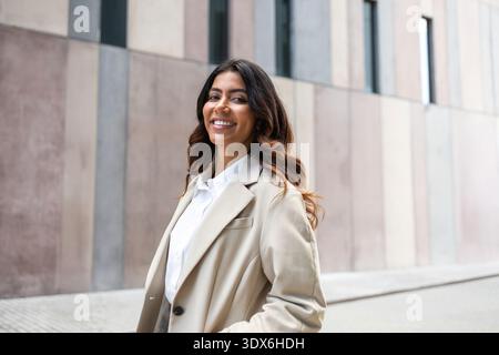 Eine hispanische Geschäftsfrau lächelt selbstbewusst, während sie in einer städtischen Umgebung steht. Sie trägt einen beigefarbenen Blazer, der ein Gefühl von Professionalität und s ausstrahlt Stockfoto