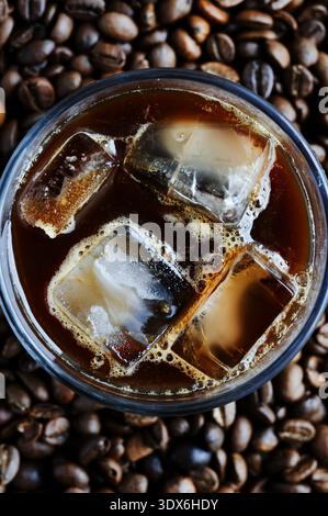 Ein Blick von oben auf ein erfrischendes Glas Eiskaffee mit großen Eiswürfeln, umgeben von dunkel gerösteten Kaffeebohnen, die einen reizvollen Kontrast bilden Stockfoto