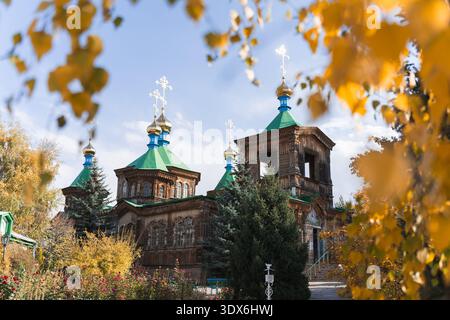Eine malerische hölzerne orthodoxe Kirche mit grünen Kuppeln und goldenen Kreuzen steht inmitten von lebhaftem Herbstlaub in Kirgisistan, das historische Religionen zeigt Stockfoto