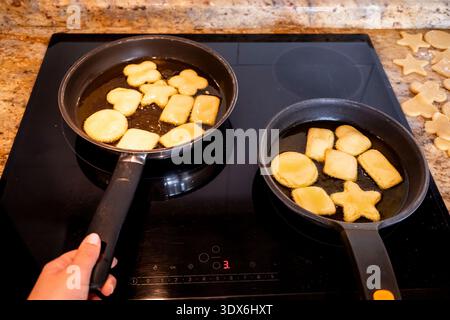 Ein Nahbild von zwei Pfannen auf einem elektrischen Herd, jede gefüllt mit einzigartig geformtem, hausgemachtem Keksenteig, der zur goldenen Perfektion gekocht wird Stockfoto