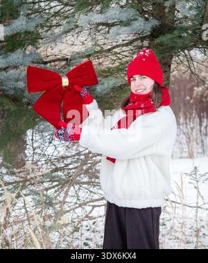 Junge Frau, die immergrünen Baum mit großer roter Schleife im schneebedeckten Park dekoriert, warme Winterkleidung trägt und in die Kamera lächelt. Stockfoto